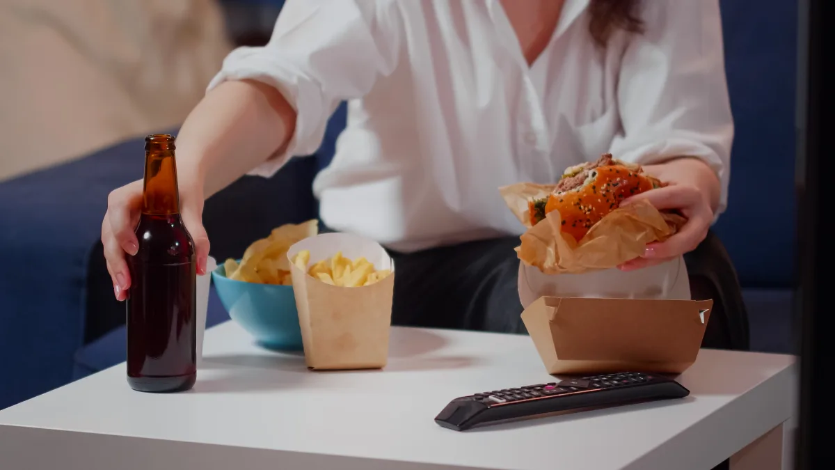 Close up of table with delivery fast food and person eating burger with bottle of beer. Hands holding hamburger while enjoying takeaway meal and sitting on sofa in living room after work
